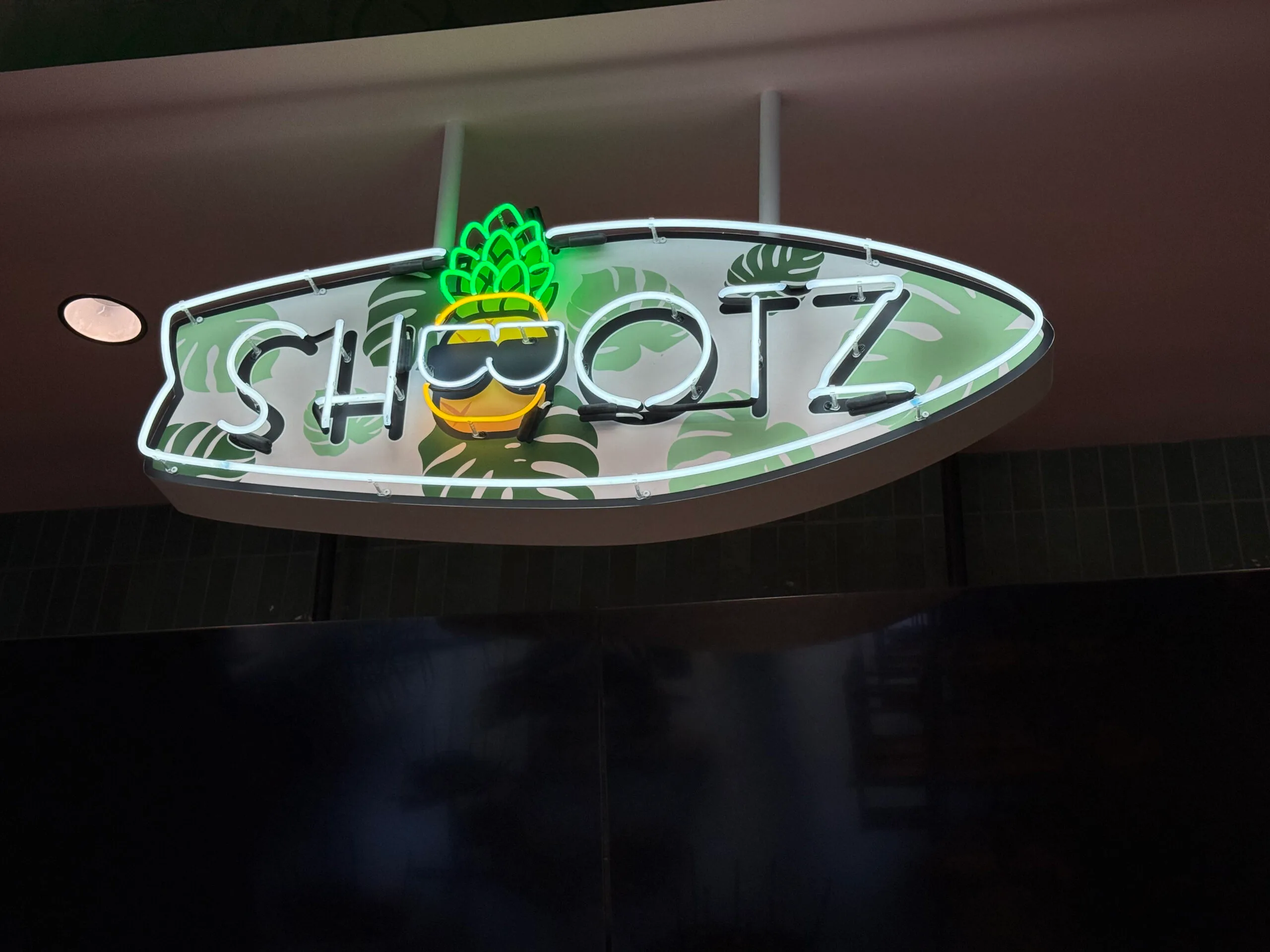 Close-up of the Shootz neon blade sign: white neon lettering and a green and yellow neon pineapple wearing sunglasses on a surfboard-shaped cabinet with a monstera-leaf print