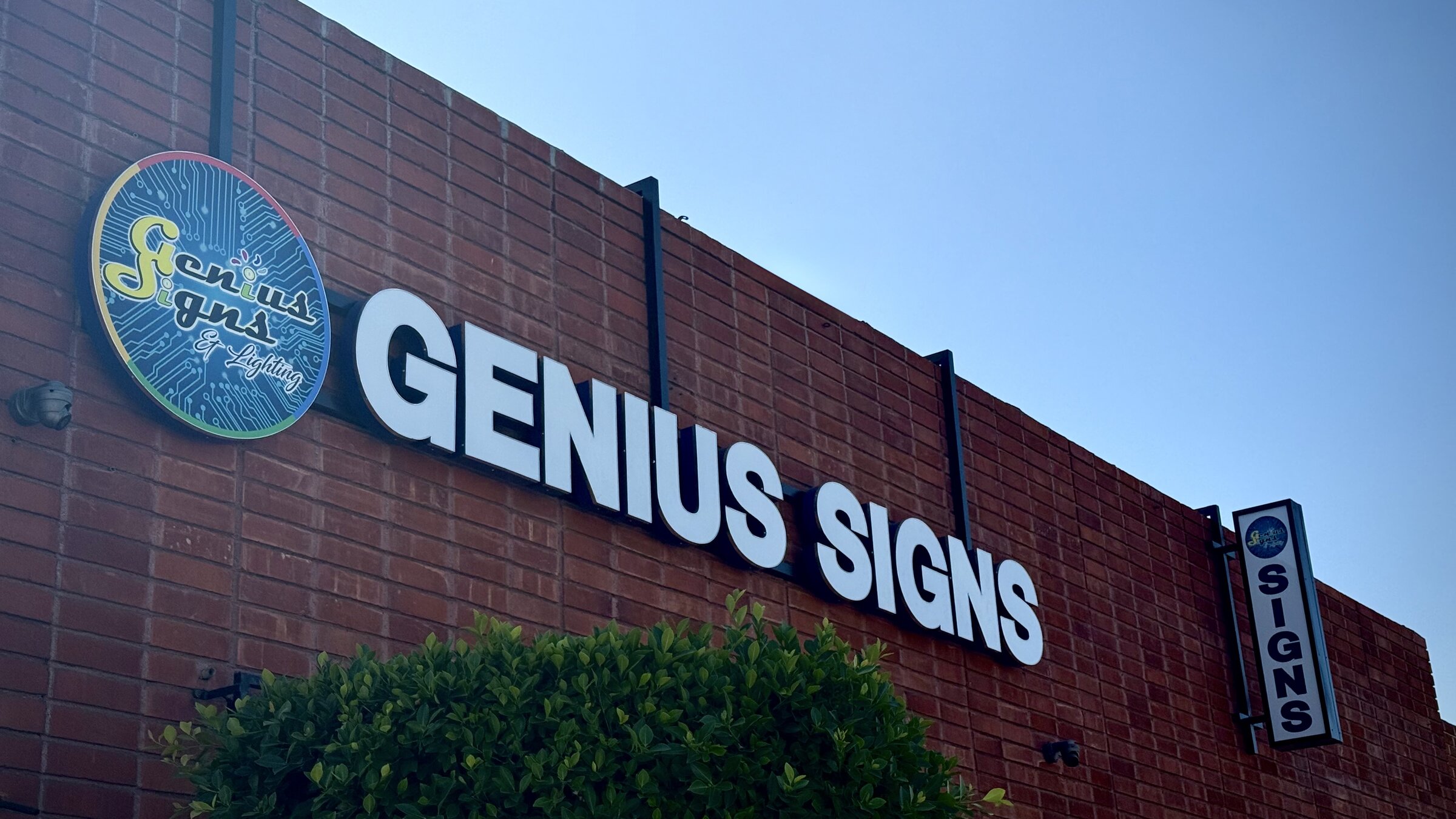 Genius Signs storefront showing illuminated front lit channel letters, circular shop logo disc, and projecting blade sign